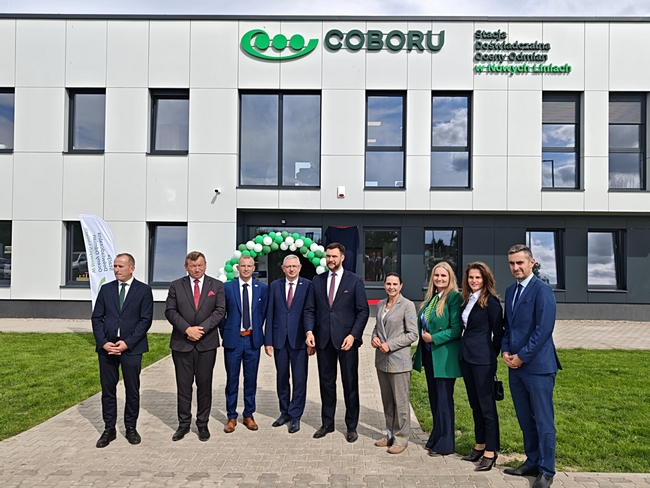 Grupa uczestników uroczystości przed nowym budynkiem SDOO Nowe Linie. W tle widoczne logo COBORU, nazwa Stacji oraz wejście udekorowane zielono-białymi balonami.