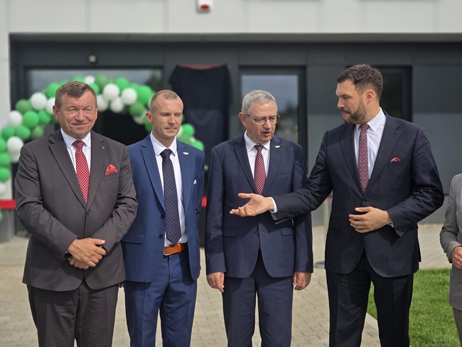 Grupa uczestników uroczystości rozmawia przed wejściem do nowego budynku SDOO Nowe Linie. W tle widoczna dekoracja z zielono-białych balonów.
